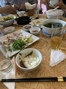 Bối cảnh là một bữa ăn tại một nhà hàng, có nhiều người đang ngồi quanh một bàn ăn. Trên bàn được trải một chiếc khăn màu vàng có hoa văn. Mọi người đang dùng đũa để ăn cơm. Bữa ăn có nhiều món đã được dọn ra, bao gồm một đĩa có thịt luộc, tôm, rau thơm và quả trông giống quả vả hoặc sung thái lát. Ngoài ra còn có một bát canh rau lớn, một nồi đất đen đựng món kho, và nhiều bát nước chấm nhỏ. Mỗi người có một bát cơm riêng. Trên bàn có các cốc thủy tinh đựng nước và một cốc đựng đồ uống màu vàng như bia hoặc trà. Có một hộp đựng khăn giấy, trên khăn giấy có in chữ "Nhà hàng" nhưng phần tên riêng không rõ.。（日本語訳）。背景はレストランでの食事で、多くの人が食卓を囲んで座っています。テーブルには模様の入った黄色いテーブルクロスが敷かれています。皆は箸を使って食事をしています。食事には多くの料理が出されており、茹で豚、エビ、香草、そしてイチジクまたはイチジクに似た果物のスライスが乗った一皿が含まれています。その他にも、大きな野菜スープの鉢、煮込み料理が入った黒い土鍋、そして多くの小さなつけだれの小鉢があります。各自にそれぞれご飯茶碗があります。テーブルの上には水が入ったガラスのコップと、ビールかお茶のような黄色い飲み物が入ったコップがあります。ティッシュペーパーの箱があり、そのティッシュペーパーには「レストラン」という文字が印刷されていますが、固有名詞の部分は不明瞭です。