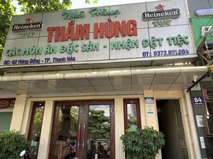 Bức ảnh chụp mặt tiền của một nhà hàng có tên 'Nhà Hàng THẮM HÙNG' tại thành phố Thanh Hóa. Các thông tin bằng chữ có thể đọc được trên biển hiệu chính bao gồm tên nhà hàng, dịch vụ 'CÁC MÓN ĂN ĐẶC SẢN - NHẬN ĐẶT TIỆC', địa chỉ 'ĐC: 62 Hàng Đồng - TP. Thanh Hóa', và số điện thoại 'ĐT: 0373.851.694'. Biển hiệu cũng có logo của thương hiệu bia 'Heineken'. Ngoài ra, có các biển số nhà '62 HÀNG ĐỒNG' và '64 HÀNG ĐỒNG'. Một tấm áp phích trên tường ghi 'NÓI LỜI HAY LÀM VIỆC TỐT HÀNH ĐỘNG THÂN THIỆN'. Khung cảnh là một nhà hàng trên phố vào ban ngày, có cây xanh ở phía trước. Lối vào là cửa kính, qua đó có thể thấy một vài khách hàng đang ngồi ăn bên trong. Một camera an ninh được lắp đặt dưới mái hiên.。（日本語訳）。タインホア市にある「THẮM HÙNGレストラン」という名前のレストランの正面を撮影した写真です。メインの看板で読み取れる文字情報には、レストラン名、サービス内容「名物料理各種 - 宴会予約受付」、住所「ĐC: 62 Hàng Đồng - TP. Thanh Hóa」、電話番号「ĐT: 0373.851.694」が含まれます。看板にはビールブランド「ハイネケン」のロゴもあります。その他に、「62 HÀNG ĐỒNG」と「64 HÀNG ĐỒNG」という番地表示があります。壁のポスターには「良い言葉を話し、良い行いをし、親切に行動しよう」と書かれています。日中の街角にあるレストランの風景で、正面には緑の木があります。入口はガラスのドアで、そこを通して中に何人かのお客さんが座って食事をしているのが見えます。軒下に防犯カメラが設置されています。