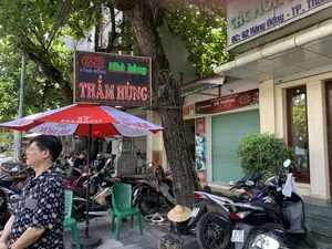 Bối cảnh là một vỉa hè trên một con phố ở thành phố Thanh Hóa. Có nhiều xe máy đang đậu san sát nhau. Một người đàn ông mặc áo sơ mi hoa văn đang đứng ở phía trước. Ở phía sau, một người phụ nữ đang đứng cạnh xe máy. Có một cây lớn trên vỉa hè, thân cây được quấn dây đèn trang trí. Một chiếc ô lớn màu đỏ và trắng của hãng bia "HABECO" (BIA HÀ NỘI) đang mở. Có một vài chiếc ghế nhựa màu xanh và một chiếc nón lá đặt trên một chiếc xe máy. Các biển hiệu có thể đọc được bao gồm: một biển hiệu đèn LED lớn cho "Nhà hàng THẮM HÙNG" ở "62 HÀNG ĐỒNG"; một biển hiệu cho "CÀ PHÊ RANG XAY NGUYÊN CHẤT CÔ HƯỜNG" ở "60 Hàng Đồng, P Điện Biên, TP Thanh Hóa", chuyên bán "TRÀ - SINH TỐ" và là của thương hiệu "TRUNG NGUYÊN INTERNATIONAL"; và một biển hiệu khác phía trên ghi "ĐC: 62 Hàng Đồng - TP. Th...".。（日本語訳）。背景はタインホア市のある通りの歩道です。たくさんのバイクが隣り合って停められています。柄物のシャツを着た男性が手前に立っています。奥では、一人の女性がバイクの隣に立っています。歩道には大きな木があり、その幹には装飾用の電飾が巻かれています。ビールブランド「HABECO」（BIA HÀ NỘI）の赤と白の大きな傘が開いています。青いプラスチックの椅子がいくつかあり、バイクの上にはノンラーが一つ置かれています。読み取れる看板には、「62 HÀNG ĐỒNG」にある「Nhà hàng THẮM HÙNG」の大きなLED看板、「60 Hàng Đồng, P Điện Biên, TP Thanh Hóa」にあり、「TRÀ - SINH TỐ」を専門に販売し、「TRUNG NGUYÊN INTERNATIONAL」ブランドのものである「CÀ PHÊ RANG XAY NGUYÊN CHẤT CÔ HƯỜNG」の看板、そしてその上にある「ĐC: 62 Hàng Đồng - TP. Th...」と書かれた別の看板が含まれます。