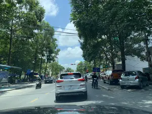 Hình ảnh ghi lại cảnh giao thông trên một con đường phố vào ban ngày, được chụp từ bên trong một chiếc ô tô. Các phương tiện như ô tô và xe máy đang dừng chờ đèn tín hiệu giao thông màu đỏ. Ở phía trước là một chiếc ô tô Hyundai Tucson màu trắng. Bên lề đường bên phải, một chiếc xe bán tải màu cam và một chiếc ô tô Toyota Yaris màu bạc đang đỗ. Hai bên đường có nhiều cây xanh lớn, tán lá rậm rạp, che bóng mát. Phía trên có hệ thống dây điện chằng chịt. Bầu trời trong xanh và có mây. Có thể đọc được nội dung trên các biển báo và biển hiệu:。- Biển chỉ đường màu xanh lam: "LÀNG HẠC 1 Km", "GA THANH HÓA 0.5 Km", "QUỐC LỘ 1A 0.5 Km".。- Biển hiệu khách sạn: "ĐẠI VIỆT HOTEL 50M", "KHU VỰC ĐỖ XE".。- Biển hiệu đèn neon màu xanh lá cây: "BODY FOOT".。（日本語訳）。昼間の街路の交通風景を車内から撮影した画像です。自動車やバイクなどの車両が赤信号で停止しています。前方には白いヒュンダイ・ツーソンがいます。右側の路肩には、オレンジ色のピックアップトラックとシルバーのトヨタ・ヤリスが駐車しています。道路の両側には大きな緑の木々が多く、葉が茂り、日陰を作っています。上には電線が張り巡らされています。空は青く、雲があります。看板や標識の内容を読み取ることができます：。- 青い道路標識：「LÀNG HẠC 1 Km」、「GA THANH HÓA 0.5 Km」、「QUỐC LỘ 1A 0.5 Km」。- ホテルの看板：「ĐẠI VIỆT HOTEL 50M」、「KHU VỰC ĐỖ XE」。- 緑色のネオンサイン：「BODY FOOT」。