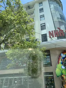 Bức ảnh chụp một tòa nhà cao tầng màu trắng với kiến trúc hiện đại, có một phần mặt tiền được làm bằng kính cong. Phía trước tòa nhà có một cây xanh lớn với tán lá rậm rạp. Trên tòa nhà có một biển hiệu lớn màu đỏ với các chữ cái "NHÀ S...", phần còn lại của chữ bị khuất. Tầng trệt của tòa nhà là các cửa hàng. Nhìn qua cửa kính, có thể thấy một cửa hàng trưng bày nhiều loại gấu bông và đồ chơi. Một cửa hàng khác dường như bán hoa giả và bình hoa. Ở phía bên phải, có một tấm biển trang trí nhiều màu sắc với hình cầu vồng, đám mây, và một con hươu cao cổ, gợi ý đây là khu vực dành cho trẻ em, có thể là một nhà sách hoặc cửa hàng đồ chơi. Bầu trời trong xanh và có nắng.。（日本語訳）。写真には、近代的な建築様式の白い高層ビルが写っており、ファサードの一部は曲面ガラスでできています。ビルの前には、葉が鬱蒼と茂った大きな緑の木があります。ビルには「NHÀ S...」という文字が書かれた大きな赤い看板があり、残りの文字は隠れています。ビルの1階は店舗になっています。ガラス窓越しに、たくさんの種類のテディベアやおもちゃを陳列している店が見えます。別の店では造花や花瓶を売っているようです。右側には、虹、雲、キリンの絵が描かれたカラフルな装飾看板があり、ここが子供向けのエリア、おそらく書店かおもちゃ屋であることを示唆しています。空は青く晴れ渡っています。