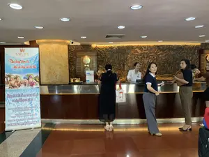 Bối cảnh là sảnh chờ của một khách sạn, tại khu vực quầy lễ tân. Một số khách hàng đang đứng trước quầy, có vẻ như đang làm thủ tục hoặc trao đổi thông tin. Một nữ nhân viên lễ tân mặc áo sơ mi trắng đang phục vụ khách. Phía sau quầy có một bức phù điêu lớn bằng gỗ chạm khắc các họa tiết truyền thống Việt Nam. Sàn nhà được lát gạch bóng.。Ở phía bên trái có một tấm biển quảng cáo dạng cuộn (standee) với các thông tin được ghi lại như sau:。SAO MAI。Thả lòng tâm tư khi ta đến với bể bơi。Chúng tôi đi bơi để xua tan sự mệt mỏi và căng thẳng sau giờ học。GIÁ VÉ CHỈ TỪ 50K (ĐỐI VỚI TRẺ EM)。70K (ĐỐI VỚI NGƯỜI LỚN)。THỜI GIAN MỞ CỬA:。6H00 - 9H00 & 15H00 - 19H00。HOTLINE: 0981.247.789 - 033.4747.666。ĐC: 20 PHAN CHU TRINH, P. ĐIỆN BIÊN, TP. THANH HÓA。（日本語訳）。背景はホテルのロビー、フロントカウンターエリアです。数名のお客様がカウンターの前に立っており、手続きをしているか、情報を交換しているようです。白いシャツを着た女性フロントスタッフがお客様に対応しています。カウンターの後ろには、ベトナムの伝統的な模様が彫られた大きな木製のレリーフがあります。床は光沢のあるタイルで敷き詰められています。左側には、以下の情報が記載されたロールアップバナー（スタンディ）があります。SAO MAI。プールに来て、心をリラックスさせましょう。私たちは授業後の疲れやストレスを解消するために泳ぎに行きます。チケット料金 わずか50Kから（子供）。70K（大人）。営業時間：。6時00分～9時00分 & 15時00分～19時00分。ホットライン：0981.247.789 - 033.4747.666。住所：タインホア市ディエンビエン区ファン・チュー・チン通り20番地