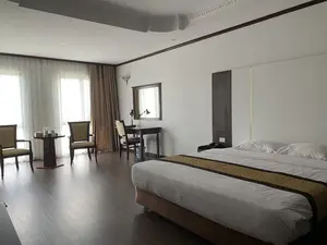 Đây là hình ảnh chụp bên trong một căn phòng khách sạn. Căn phòng được bài trí gọn gàng và sạch sẽ. Ở phía bên phải là một chiếc giường đôi lớn được trải ga trắng tinh, có hai chiếc gối và một dải vải trang trí màu vàng nâu chạy dọc giữa giường. Sát tường là bàn làm việc bằng gỗ với một chiếc ghế, một chiếc gương và đèn bàn. Gần cửa sổ lớn là một bộ bàn tròn nhỏ và hai chiếc ghế, trên bàn có đặt hai chai nước và hộp khăn giấy. Cửa sổ có hai lớp rèm, một lớp mỏng màu trắng và một lớp dày màu nâu. Sàn nhà được lát gỗ màu tối. Không có người hay hoạt động nào diễn ra trong phòng. Không có bất kỳ chữ viết nào có thể nhìn thấy.。（日本語訳）。これはホテルの客室の内部を撮影した写真です。部屋はきちんと整頓され、清潔です。右側には、真っ白なシーツが敷かれた大きなダブルベッドがあり、枕が2つと、ベッドの中央を縦に走る黄褐色の装飾布があります。壁際には、椅子、鏡、電気スタンドが備わった木製の机があります。大きな窓の近くには、小さな円卓と椅子2脚のセットがあり、テーブルの上には水が2本とティッシュボックスが置かれています。窓には2層のカーテンがあり、1層は薄い白色、もう1層は厚い茶色です。床は暗い色のフローリングです。部屋の中には人や活動はありません。