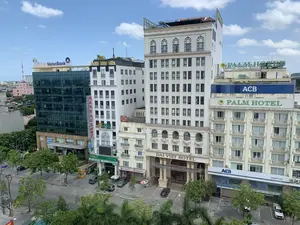 Bức ảnh chụp từ trên cao một góc phố đô thị tại Việt Nam, có thể là thành phố Thanh Hóa, vào một ngày trời trong xanh có mây. Khung cảnh nổi bật với nhiều tòa nhà cao tầng san sát nhau, là trụ sở của các khách sạn, ngân hàng và dịch vụ. Các biển hiệu có thể nhận dạng bao gồm: 'DAI VIET HOTEL', 'PALM HOTEL', 'VietinBank' trên một tòa nhà có mặt tiền bằng kính, 'HOA SEN SPA' với chữ màu vàng. Ngoài ra, có rất nhiều chi nhánh ngân hàng như 'ACB' (Ngân hàng Á Châu), 'OCB' (Ngân hàng Phương Đông) với dòng chữ 'CHUYỂN TIỀN QUỐC TẾ', và 'SeABank'. Một biển hiệu dọc màu đỏ có chữ 'MASSAGE' cũng có thể được nhìn thấy. Dưới đường, có một vài chiếc ô tô đang đỗ trong bãi đậu xe. Cây xanh được trồng dọc theo đường phố. Không có hoạt động đông đúc của người đi bộ, cho thấy một khoảnh khắc tương đối yên tĩnh trên một con phố thương mại.。（日本語訳）。ベトナムの都市の街角を上空から撮影した写真で、おそらくタインホア市だと思われます。晴れた青空に雲が浮かぶ日です。多くの高層ビルが隣接して建ち並ぶ光景が際立っており、ホテル、銀行、サービスの拠点となっています。認識できる看板には、「DAI VIET HOTEL」、「PALM HOTEL」、ガラス張りのファサードを持つ建物にある「VietinBank」、黄色い文字の「HOA SEN SPA」などがあります。その他にも、「ACB」（アジア商業銀行）、「OCB」（フンドン銀行）には「CHUYỂN TIỀN QUỐC TẾ（国際送金）」の文字があり、「SeABank」など、多くの銀行支店があります。「MASSAGE」と書かれた赤い縦長の看板も見えます。下の道路では、数台の車が駐車場に停まっています。街路に沿って緑の木々が植えられています。歩行者の賑やかな活動は見られず、商業地区の通りが比較的静かな瞬間であることを示しています。