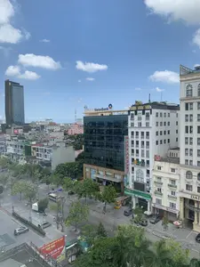 Bức ảnh chụp một góc phố của thành phố Thanh Hóa từ trên cao vào một ngày trời trong, mây trắng. Con đường rộng rãi, có nhiều cây xanh, với một vài chiếc ô tô và xe máy đang lưu thông và đậu ven đường. Cảnh quan đô thị bao gồm nhiều tòa nhà cao tầng hiện đại. Nổi bật là các biển hiệu của nhiều doanh nghiệp: tòa nhà văn phòng bằng kính có logo và chữ 'VietinBank' trên nóc; một tòa nhà màu trắng với biển hiệu 'HOA SEN SPA' màu vàng; các chi nhánh ngân hàng 'OCB' (Ngân Hàng Phương Đông) và 'SeABank'; một biển hiệu dọc ghi chữ 'MASSAGE'. Ở phía xa là một tòa nhà chọc trời. Phía dưới bên trái có một tấm biển quảng cáo lớn màu đỏ với nội dung: 'TÂM TỔ CHỨC SỰ KIỆN VÀ ẨM THỰC SAO MAI', '...TIỆC CƯỚI, HỘI NGHỊ CHUYÊN NGHIỆP ...nhất tại Thanh Hóa'.。（日本語訳）。晴れた白い雲の日に、上空から撮影されたタインホア市の一角の写真。道は広く、緑の木々が多く、数台の自動車やバイクが通行したり、道端に駐車したりしている。都市景観には、多くの近代的な高層ビルが含まれている。目立っているのは多くの企業の看板である：屋上にロゴと「VietinBank」の文字があるガラス張りのオフィスビル、黄色い「HOA SEN SPA」の看板がある白い建物、銀行支店の「OCB」（フンドン銀行）と「SeABank」、そして「MASSAGE」と書かれた縦長の看板。左下には、「TÂM TỔ CHỨC SỰ KIỆN VÀ ẨM THỰC SAO MAI」（サオマイ・イベント・飲食センター）、「...TIỆC CƯỚI, HỘI NGHỊ CHUYÊN NGHIỆP ...nhất tại Thanh Hóa」（...タインホアで最もプロフェッショナルな結婚披露宴、会議...）という内容の大きな赤い広告看板がある。