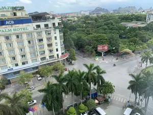 Đây là quang cảnh một ngã tư đường phố tại một thành phố, được chụp từ trên cao. Khung cảnh bao gồm nhiều tòa nhà, cây xanh và những dãy núi ở phía xa. Hoạt động chính là giao thông đô thị với một vài ô tô và xe máy đang di chuyển trên đường. Một người đang đi bộ trên vỉa hè.。Các văn bản và chữ viết có thể nhận dạng được bao gồm:。- Trên tòa nhà cao tầng bên trái: "PALM HOTEL", "ACB", "NGÂN HÀNG Á CHÂU", "Tỷ sở Ngân hàng Á Châu - Chi nhánh Thanh...", và "Welcome to Palm Hotel".。- Trên một tòa nhà khác: "THANH GROUP".。- Trên một tòa nhà ở xa: "DAI VIET PALACE".。- Trên một biển hiệu ở góc đường: "NHA SĨ..." (phần còn lại bị che khuất).。- Biển quảng cáo lớn tại ngã tư: "GENERALI", "TẬP ĐOÀN BẢO HIỂM SỐ 1 TẠI Ý", "#SốngNhưÝ".。- Một biển hiệu nhỏ hơn có chữ "MASSAGE".。Các vật thể đáng chú ý khác bao gồm một cổng chào trang trí bắc qua đường, nhiều cây cọ và cây xanh dọc vỉa hè, các cột cờ treo quốc kỳ Việt Nam (cờ đỏ sao vàng) và các lá cờ khác ở phía trước.。（日本語訳）。これは、ある都市の街の交差点の光景で、上空から撮影されたものです。風景には、多くの建物、緑の木々、そして遠くの山脈が含まれています。主な活動は都市交通で、数台の自動車とバイクが道路を移動しています。一人の人が歩道を歩いています。認識可能なテキストと文字には、以下が含まれます。- 左側の高層ビルには：「PALM HOTEL」、「ACB」、「NGÂN HÀNG Á CHÂU」（アジア商業銀行）、「Tỷ sở Ngân hàng Á Châu - Chi nhánh Thanh...」（アジア商業銀行本店 - タイン支店...）、そして「Welcome to Palm Hotel」。- 別の建物には：「THANH GROUP」。- 遠くの建物には：「DAI VIET PALACE」。- 角の看板には：「NHA SĨ...」（歯科医...）（残りの部分は隠れています）。- 交差点の大きな広告看板には：「GENERALI」、「TẬP ĐOÀN BẢO HIỂM SỐ 1 TẠI Ý」（イタリアNo.1の保険グループ）、「#SốngNhưÝ」。- 「MASSAGE」という文字がある小さな看板。その他の注目すべき物体には、道路をまたぐ装飾的な歓迎ゲート、歩道沿いの多くのヤシの木と緑の木々、手前にベトナム国旗（金星紅旗）やその他の旗を掲げた旗竿が含まれます。