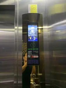 Đây là hình ảnh chụp bên trong một thang máy có vách làm bằng inox sáng bóng. Trọng tâm của bức ảnh là bảng điều khiển thang máy. Trên bảng điều khiển có một màn hình LCD hiển thị ngày 07/07/2024, giờ 13:21, và cho biết thang máy đang ở tầng 2. Bên dưới màn hình là các thông tin về thang máy: tên công ty "THANG MÁY MIỀN TRUNG", sức chứa "CAPACITY: 10 PERSONS", tải trọng "LOAD: 750 KG", biển báo "NO SMOKING" (Cấm hút thuốc) và số điện thoại "HOTLINE: 19000138". Phía trên màn hình có dán một "TEM KIỂM ĐỊNH" màu vàng. Tem ghi rõ thông tin của đơn vị kiểm định là "CÔNG TY TNHH MTV KIỂM ĐỊNH KTAT VIỆT NAM" có địa chỉ tại Hà Nội, tên đối tượng là "THANG MÁY ĐIỆN (P2)", ngày kiểm định là 08/12/2023 và có hiệu lực đến 08/11/2024. Bên dưới là các nút bấm chọn tầng và nút chuông báo động. Có hình ảnh phản chiếu của một người đàn ông đeo kính trên bề mặt kim loại của bảng điều khiển.。（日本語訳）。これは、光沢のあるステンレス鋼製の壁を持つエレベーターの内部で撮影された写真です。写真の中心はエレベーターの操作盤です。操作盤には、2024年7月7日、13時21分という日時を表示し、エレベーターが2階にいることを示すLCD画面があります。画面の下には、エレベーターに関する情報があります：会社名「THANG MÁY MIỀN TRUNG」、定員「CAPACITY: 10 PERSONS」、積載量「LOAD: 750 KG」、標識「NO SMOKING」（禁煙）、そして電話番号「HOTLINE: 19000138」。画面の上には、黄色の「検査済証」が貼られています。その証票には、検査機関の情報が明記されています。検査機関はハノイに住所を置く「CÔNG TY TNHH MTV KIỂM ĐỊNH KTAT VIỆT NAM」、対象名は「THANG MÁY ĐIỆN (P2)」、検査日は2023年12月8日で、有効期限は2024年11月8日までです。その下には、階数選択ボタンと非常ベルボタンがあります。操作盤の金属面に、眼鏡をかけた男性の姿が映り込んでいます。