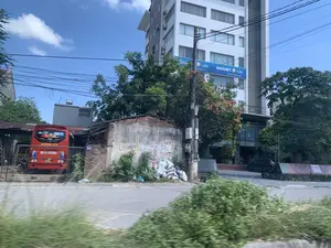 Đây là hình ảnh chụp một con đường tại một đô thị, có thể là ở thành phố Thanh Hóa, dưới bầu trời xanh và nắng. Khung cảnh cho thấy sự tương phản giữa một tòa nhà văn phòng hiện đại, cao tầng màu trắng và một công trình cũ kỹ, có thể là nhà kho hoặc nhà ở, đã xuống cấp nằm bên cạnh.。Trên tòa nhà hiện đại có biển hiệu lớn màu xanh với dòng chữ 'BAOVIET Life' và 'BẢO VIỆT NHÂN THỌ'. Trước tòa nhà có một chiếc ô tô màu đen đang đỗ.。Công trình cũ có tường gạch và bê tông, trên tường có nhiều dòng chữ quảng cáo được sơn đã phai màu, có thể đọc được một phần như 'LÀM LỢP...'. Dựa vào tường là một đống bao tải.。Ở bên trái, phía sau hàng rào, một chiếc xe khách màu đỏ của nhà xe 'DŨNG VY' đang đỗ. Trên xe có chữ 'UNIVERSE'.。Các chi tiết khác trong ảnh bao gồm cây xanh, một cột điện với hệ thống dây điện và công tơ chằng chịt, và một con đường nhựa ở phía trước.。（日本語訳）。これは、晴れた青空の下、ある都市の、おそらくはタインホア市の一本の道を撮影した写真です。その光景は、近代的な高層の白いオフィスビルと、その隣にある、倉庫か住居と思われる老朽化した古い建物との対比を示しています。近代的なビルには、「BAOVIET Life」と「BẢO VIỆT NHÂN THỌ」という文字が書かれた大きな青い看板があります。ビルの前には黒い車が一台駐車しています。古い建物はレンガとコンクリートの壁で、壁には色褪せた塗装の広告文がいくつも書かれており、「LÀM LỢP...」のような一部を読むことができます。壁に寄りかかって、麻袋の山があります。左側、柵の後ろには、「DŨNG VY」社の赤いバスが一台駐車しています。バスには「UNIVERSE」という文字があります。写真のその他の詳細には、緑の木々、複雑に絡み合った電線とメーターが付いた電柱、そして手前のアスファルトの道路が含まれます。