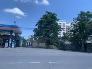 Bản ghi văn bản từ hình ảnh: Trên biển hiệu lớn màu xanh của trạm xăng có ghi: 'SƠN PHÁT PETRO - THƯƠNG NHÂN XUẤT NHẬP KHẨU XĂNG DẦU', 'CÔNG TY CỔ PHẦN ĐẦU TƯ XD & TM MINH HƯƠNG', và tên trạm xăng 'XĂNG DẦU MINH HƯƠNG' cùng số điện thoại 'ĐT: 02373.941.293'. Trên các cột bơm xăng có hiển thị giá: 'DIESEL 0.05S-II' là 21.590 và 'XĂNG RON 95-III' là 24.020, với logo 'AP PETRO'.。Mô tả cảnh quan và hoạt động: Hình ảnh chụp một con đường nhựa trong một khu đô thị vào một ngày trời trong xanh, có nắng. Địa điểm chính trong ảnh là một trạm xăng dầu ('cây xăng') tên là 'Xăng dầu Minh Hương' nằm ở bên trái. Có hai cột bơm xăng và một vài chiếc xe máy đang đỗ gần đó. Ở giữa và phía sau trạm xăng là những cây xanh lớn. Phía sau hàng cây là một tòa nhà chung cư hiện đại, cao tầng, sơn màu trắng. Xen kẽ là một vài ngôi nhà cấp bốn cũ hơn và những bức tường đá. Bầu trời quang đãng với vài gợn mây trắng. Không có hoạt động mua bán hay giao thông tấp nập nào được ghi lại, khung cảnh khá yên tĩnh.。（日本語訳）。画像からのテキスト書き起こし：ガソリンスタンドの大きな青い看板には、「SƠN PHÁT PETRO - 石油輸出入業者」、「MINH HƯƠNG建設投資貿易株式会社」、そしてガソリンスタンド名「XĂNG DẦU MINH HƯƠNG」と電話番号「電話：02373.941.293」が記載されています。給油機には、「DIESEL 0.05S-II」が21,590、「XĂNG RON 95-III」が24,020という価格が「AP PETRO」のロゴと共に表示されています。風景と活動の説明：画像は、晴れた青空の日に、都市部のアスファルト道路を撮影したものです。写真の主な場所は、左側にある「Xăng dầu Minh Hương」という名前のガソリンスタンド（「cây xăng」）です。2つの給油機と、その近くに数台のバイクが停まっています。ガソリンスタンドの中央と後方には大きな緑の木々があります。並木の背後には、白く塗られた現代的な高層マンションがあります。その間に、いくつかの古い平屋建ての家と石垣が点在しています。空は晴れ渡り、白い雲がいくつかあります。活発な売買や交通の往来は記録されておらず、景色はかなり静かです。