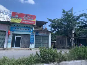 Bối cảnh là một con đường ở thành phố Thanh Hóa vào một ngày nắng, trời trong xanh. Bên đường là một dãy các cửa hàng liền kề, nhiều cửa hàng trong số đó đang đóng cửa bằng cửa cuốn kim loại.。**Chữ viết được ghi lại từ các biển hiệu:**。1.  **Biển quảng cáo lớn (Vietjet Air):**。*   `VietjetAir.com`。*   `PHÒNG VÉ ĐỨC HOA`。*   `Số nhà 42, Tổ 9, Phố Bắc Sơn, Phường An Hưng, Thành Phố Thanh Hóa`。*   `Hotline: 0399 842 602 - 0914 745 436`。*   `Email: leminhduc36n1@gmail.com`。2.  **Biển hiệu tiệm cắt tóc (màu xanh dương):**。*   `Minh Đức - Từ 2010`。*   `CẮT TÓC NAM`。*   `Lấy Ráy Tai Kỹ Thuật - Gội Nhuộm Thời Trang`。*   `ĐT: 0355 648 496 - 091 47 45 436`。*   Trên cửa có chữ `Mr. Minh Đức` và số nhà `42`.。3.  **Biển hiệu dọc (bán vé máy bay):**。*   `ĐIỂM BÁN VÉ MÁY BAY`。*   `TẤT CẢ CÁC CHẶNG NỘI ĐỊA - QUỐC TẾ`。*   Logo của VietjetAir, Jetstar。*   `www.abay.vn`, `Hotline 0399 842 602`。4.  **Biển hiệu cửa hàng bia mộ (màu trắng):**。*   `BIA MỘ ĐÁ`。*   `Thiệu Cường`。*   `40/9 BẮC SƠN`。*   `ẢNH MEN SỨ - BẮN CÁT ĐÁ GRANIT`。*   `ĐT: 0974 791 266`。5.  **Biển hiệu cửa hàng bên trái (màu hồng/tím, một phần bị che):**。*   `HUẾ HÀ`。*   `SÀO CHỊU LỰC`, `RÈM CỬA`, `CUNG CẤP PHỤ KIỆN`, `SỬA`, `IN ÁO BỘ`。6.  **Biển tròn nhỏ màu đỏ:** `Vietlott`。**Mô tả cảnh vật và hoạt động:**。*   **Loại địa điểm:** Đây là một con phố thương mại ở thành phố Thanh Hóa. Các tòa nhà là những cửa hàng mặt tiền điển hình ở Việt Nam.。*   **Hoạt động:** Không có hoạt động rõ rệt nào của con người. Hầu hết các cửa hàng đều đóng cửa, cho thấy đây có thể là thời gian nghỉ trưa hoặc một ngày nghỉ. Con đường phía trước vắng vẻ.。*   **Vật thể:** Các cửa hàng với cửa cuốn kim loại, một biển quảng cáo lớn của hãng hàng không, nhiều biển hiệu nhỏ hơn, một con đường nhựa, cây xanh, và một cột điện với hệ thống dây điện phức tạp.。（日本語訳）。背景は、晴れた日のタインホア市の通りで、空は青く澄んでいます。道端には店舗が軒を連ねており、その多くは金属製のシャッターで閉まっています。**看板から記録された文字：**。1.  **大きな広告看板（ベトジェットエア）：**。*   `VietjetAir.com`。*   `ドゥックホア航空券販売所`。*   `タインホア市アンフン坊バクソン通り9組42番地`。*   `ホットライン: 0399 842 602 - 0914 745 436`。*   `Eメール: leminhduc36n1@gmail.com`。2.  **理髪店の看板（青色）：**。*   `ミン・ドゥック - 2010年から`。*   `男性カット`。*   `匠の耳かき - おしゃれシャンプー・カラー`。*   `電話: 0355 648 496 - 091 47 45 436`。*   ドアには「Mr. Minh Đức」の文字と番地「42」がある。3.  **縦長の看板（航空券販売）：**。*   `航空券販売所`。*   `国内線・国際線全路線`。*   ベトジェットエア、ジェットスターのロゴ。*   `www.abay.vn`, `ホットライン 0399 842 602`。4.  **墓石店の看板（白色）：**。*   `石の墓石`。*   `ティエウ・クオン`。*   `バクソン 40/9`。*   `陶磁器写真 - 御影石サンドブラスト`。*   `電話: 0974 791 266`。5.  **左側の店の看板（ピンク/紫色、一部が隠れている）：**。*   `フエ・ハ`。*   `高耐久物干し竿`, `カーテン`, `付属品提供`, `修理`, `セット服プリント`。6.  **赤い小さな丸い看板：** `Vietlott`。**風景と活動の説明：**。*   **場所の種類：** ここはタインホア市の商店街です。建物はベトナムの典型的な店舗兼住宅です。*   **活動：** 人々の明確な活動は見られません。ほとんどの店が閉まっており、昼休みか休日である可能性を示唆しています。前の道は閑散としています。*   **物体：** 金属製のシャッターがある店、航空会社の大きな広告看板、多数の小さな看板、アスファルトの道路、緑の木々、そして複雑な配線を持つ電柱。