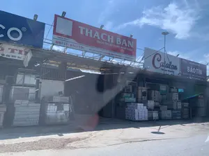 Đây là hình ảnh chụp mặt tiền của một cửa hàng vật liệu xây dựng có tên là Đại lý Bảo Tín, nằm ven đường. Phía trên cửa hàng có nhiều biển hiệu quảng cáo lớn. Biển hiệu chính, màu đỏ, ghi rõ thông tin: 'THACH BAN - GẠCH ỐP LÁT CÔNG NGHỆ ITALIA', 'SINCE 1959', 'ĐẠI LÝ: BẢO TÍN', và địa chỉ 'ĐC: XÃ THỌ TIẾN, HUYỆN TRIỆU SƠN, THANH HOÁ - ĐT: 0982.653.888'. Bên cạnh là các biển hiệu khác cho thương hiệu 'Catalan' và một biển hiệu của 'NHÀ PHÂN PHỐI BẢO TÍN' quảng cáo 'GẠCH ỐP LÁT, THIẾT BỊ VỆ SINH CAO CẤP, SƠN NỘI NGOẠI THẤT'. Phía trước và bên trong cửa hàng, nhiều chồng gạch men được đóng thùng và xếp trên các pallet. Cửa hàng có kiến trúc nhà kho với mái tôn. Bối cảnh là một ngày nắng đẹp, trời trong xanh và có mây trắng. Không có hoạt động mua bán nào được nhìn thấy rõ, chỉ có hàng hóa được trưng bày.。（日本語訳）。これは、道端にある「バオティン代理店」という名前の建材店の正面を撮影した写真です。店の上部には多くの大きな広告看板があります。赤色のメイン看板には、「THACH BAN - イタリア技術のタイル」、「SINCE 1959」、「代理店：バオティン」、そして住所「住所：タインホア省、チウソン県、トティエン社 - 電話：0982.653.888」という情報が明記されています。その隣には、「Catalan」ブランドの他の看板や、「バオティン販売代理店」の看板があり、「タイル、高級衛生陶器、内外装用塗料」を宣伝しています。店の前と内部には、箱詰めされパレットに積まれた多くのセラミックタイルの山があります。店はトタン屋根の倉庫のような建築様式です。背景は晴れた美しい日で、空は青く澄み渡り、白い雲が浮かんでいます。売買の様子ははっきりと見えず、商品が陳列されているだけです。