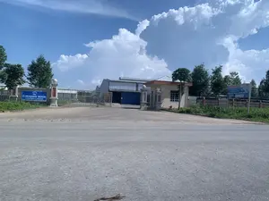 Bức ảnh chụp lối vào của một nhà máy từ phía bên kia một con đường nhựa. Đây là một nhà máy chế biến gỗ, có cổng sắt lớn đang đóng và một nhà bảo vệ bên cạnh. Phía sau cổng là các tòa nhà xưởng lớn. Bầu trời trong xanh và có nhiều mây. Xung quanh khu vực lối vào có trồng cây xanh và hàng rào.。Các văn bản trên biển hiệu có thể được ghi lại như sau:。- Biển hiệu màu xanh ở cổng bên trái: "C-HOLDING WOOD COMPANY LIMITED".。- Biển hiệu lớn ở bên phải: "NHÀ MÁY CHẾ BIẾN GỖ XUẤT KHẨU", kèm theo thông tin tuyển dụng với mức lương từ 7.000.000 đến 15.000.000 đồng.。- Biển hiệu nhỏ hơn ở dưới: "ĐIỂM THU MUA KEO CỌC - KEO GIẤY".。Không có hoạt động nào của con người được quan sát thấy trong ảnh.。（日本語訳）。写真はアスファルト道路の向こう側から工場の入り口を撮影したものです。これは木材加工工場で、大きな鉄の門が閉まっており、その隣に警備員室があります。門の後ろには大きな工場の建物があります。空は青く澄んでおり、雲が多いです。入り口の周辺には緑の木々とフェンスが植えられています。看板の文言は以下のように記録できます。- 左の門にある青い看板：「C-HOLDING WOOD COMPANY LIMITED」。- 右側の大きな看板：「輸出用木材加工工場」、給与7,000,000ドンから15,000,000ドンの求人情報付き。- その下の小さな看板：「アカシア杭 - 製紙用アカシアの買取所」。写真には人間の活動は見られません。