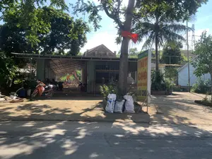 Hình ảnh ghi lại một khung cảnh ven đường ở một khu vực nông thôn. Nổi bật là một tấm biển hiệu màu vàng dựng bên đường, trên đó có dòng chữ màu đỏ "THỊT CHÓ" và "LẨU CHÓ", cùng với tên quán có thể là "QUÁN QUÝ TƯỚC". Điều này cho thấy đây là một quán ăn chuyên về các món thịt chó.。Cạnh biển hiệu là một ngôi nhà một tầng có mái hiên rộng lợp tôn. Dưới mái hiên, có một chiếc máy xúc nhỏ màu cam đang đỗ, và có thể thấy một người đang đội nón lá ngồi bên trong. Phía trước có một chiếc xe máy màu cam và một vài chiếc xe máy khác. Một lá cờ đỏ sao vàng của Việt Nam được treo trên một thân cây. Ven đường có một vài bao tải lớn. Khung cảnh xung quanh có nhiều cây xanh, bao gồm cả cây dừa, và ở phía xa có một cột điện cao thế.。（日本語訳）。画像は、ある田舎の地域の道端の光景を捉えている。道端に立てられた黄色い看板が際立っており、その上には赤い文字で「THỊT CHÓ」（犬肉）と「LẨU CHÓ」（犬鍋）、そして店名と思われる「QUÁN QUÝ TƯỚC」という文字がある。このことから、ここが犬肉料理を専門とする飲食店であることがわかる。看板の隣には、トタンで葺かれた広い軒のある一階建ての家がある。前方にはオレンジ色のバイク一台と、他のバイクが数台ある。道端には大きな袋がいくつかある。周囲の風景にはヤシの木を含む多くの緑の木があり、遠くには高圧電線の鉄塔が一本ある。