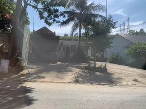 Bức ảnh chụp một khung cảnh ven đường tại một khu dân cư, có thể là ở vùng nông thôn hoặc ngoại ô. Phía trước là một con đường nhựa, phía sau là một khoảng sân đất rộng dẫn vào các ngôi nhà. Có thể thấy một vài ngôi nhà với kiến trúc khác nhau; một ngôi nhà có tường gạch, cổng sắt và một chiếc xe máy đang đỗ bên trong. Một ngôi nhà khác được sơn màu sáng với mái ngói. Giữa các ngôi nhà có nhiều cây cối xanh tươi, bao gồm một cây dừa ở trung tâm và các cây lớn khác tạo bóng mát. Ở giữa sân có một bồn cây nhỏ. Phía trước một ngôi nhà có một lò hóa vàng mã. Bên trái, có một tấm biển hiệu dựng đứng nhưng không thể đọc rõ nội dung. Gần đó có một bao tải màu trắng. Trên nền trời xanh, có thể thấy các đường dây điện và những cột điện cao thế ở phía xa. Khung cảnh yên tĩnh dưới trời nắng, không có hoạt động nào của con người được ghi lại.。（日本語訳）。写真は、ある住宅地の道端の風景を写しており、田舎か郊外の可能性があります。手前にはアスファルトの道があり、その後ろには家々へと続く広い土の庭が広がっています。異なる建築様式の家がいくつか見えます。ある家はレンガの壁と鉄の門があり、中にはバイクが一台停まっています。別の家は明るい色で塗られ、瓦屋根です。家々の間にはたくさんの青々とした木々があり、中央のココナッツの木一本と、木陰を作る他の大きな木々が含まれています。庭の中央には小さな植え込みがあります。ある家の前には、冥銭を燃やすためのかまどがあります。左側には立て看板がありますが、その内容ははっきりと読み取れません。その近くには白い袋が一つあります。青空を背景に、電線と遠くの高圧電線の鉄塔が見えます。晴れた空の下の静かな光景で、人の活動は記録されていません。