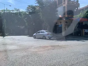 Bức ảnh chụp một góc phố tại Việt Nam vào một ngày nắng. Một chiếc xe taxi màu bạc đang đỗ ở một ngã ba đường. Bên phải là một ngôi nhà nhiều tầng, phía trước có gắn biển tên đường màu xanh ghi "ĐƯỜNG TRỊNH THỊ NGỌC LỮ". Ngôi nhà có cửa cuốn bằng kim loại đang đóng. Xung quanh có nhiều cây xanh và phía trên là bầu trời trong xanh. Con đường được trải nhựa. Khung cảnh khá yên tĩnh, không có hoạt động nào đáng chú ý diễn ra. Hình ảnh có một chút lóa sáng và phản chiếu, cho thấy nó có thể được chụp từ bên trong một chiếc xe hơi.。（日本語訳）。写真は、晴れた日のベトナムのある街角を写したものです。銀色のタクシーが1台、三叉路に停車しています。右側には多層階の家があり、その正面には「ĐƯỜNG TRỊNH THỊ NGỌC LỮ」と書かれた青い道路標識が取り付けられています。その家には金属製のシャッターがあり、閉まっています。周囲には多くの緑の木々があり、上には澄んだ青空が広がっています。道路はアスファルトで舗装されています。風景はかなり静かで、特筆すべき活動は何も行われていません。画像には少し光のまぶしさや反射があり、車内から撮影された可能性を示唆しています。