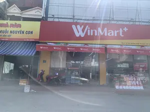 Bức ảnh chụp mặt tiền của một cửa hàng tiện lợi WinMart+ và một cửa hàng bán đặc sản nằm liền kề trên một con phố.。**Chữ viết được ghi lại:**。1.  **Biển hiệu WinMart+ (màu đỏ):** "WinMart+", khẩu hiệu "TƯƠI NGON THƯỢNG HẠNG", và địa chỉ "Khu 3, Thị Trấn Lam Sơn, Huyện Thọ Xuân, Tỉnh Thanh Hóa".。2.  **Biển hiệu Nhà Mắm (màu vàng):** "NHÀ MẮM MUỐI NGUYÊN CON", "SẦN SẬT", "- Thanh Hóa", và số điện thoại "0967.658.331".。3.  **Biển số nhà (màu xanh):** "52 LÊ THÁI TÔNG".。4.  **Thông tin khác:** Trên cửa kính có các thông báo như giờ mở cửa "6h-22h". Phía trước có trưng bày các sản phẩm như mì gói "3 Miền" và các mặt hàng khác với bảng giá.。**Mô tả cảnh vật và hoạt động:**。*   **Loại địa điểm:** Đây là mặt tiền của các cửa hàng bán lẻ trên một con phố, bao gồm một siêu thị mini (cửa hàng tiện lợi) và một cửa hàng bán đặc sản địa phương (mắm).。*   **Hoạt động cụ thể:** Khung cảnh tĩnh, không có hoạt động mua bán rõ rệt. Một chiếc xe máy đang đỗ phía trước, có thể là của khách hàng hoặc nhân viên. Hàng hóa được trưng bày bên ngoài cửa hàng để bán.。*   **Các vật thể trong ảnh:** Có hai tòa nhà với biển hiệu lớn. Một chiếc xe máy màu đỏ đang đỗ trên vỉa hè. Nhiều loại hàng hóa được trưng bày, đáng chú ý là các thùng nước khoáng "LaVie" và các thùng mì ăn liền "3 Miền".。（日本語訳）。写真は、ある通りに隣接して建つWinMart+コンビニエンスストアと特産品店の正面を撮影したものです。**記録された文字：**。1.  **WinMart+の看板（赤色）：**「WinMart+」、スローガン「TƯƠI NGON THƯỢNG HẠNG（新鮮 美味 上質）」、および住所「Khu 3, Thị Trấn Lam Sơn, Huyện Thọ Xuân, Tỉnh Thanh Hóa（タインホア省トースアン県ラムソン町第3区）」。2.  **ニャーマム（マムの家）の看板（黄色）：**「NHÀ MẮM MUỐI NGUYÊN CON（丸ごと塩漬けマムの家）」、「SẦN SẬT（コリコリ）」、「- Thanh Hóa（- タインホア）」、および電話番号「0967.658.331」。3.  **家屋番号プレート（青色）：**「52 LÊ THÁI TÔNG（レ・タイ・トン通り52番地）」。4.  **その他の情報：**ガラスドアには営業時間「6h-22h（6時～22時）」などの告知があります。店の前には「3 Miền」のインスタントラーメンなどの商品や、価格表付きのその他の品物が陳列されています。**風景と活動の描写：**。*   **場所の種類：**これはある通りにある小売店の正面であり、ミニスーパー（コンビニエンスストア）と地元の特産品（マム）を販売する店が含まれています。*   **具体的な活動：**静かな光景で、明確な売買活動は見られません。一台のバイクが前に停まっており、客または従業員のものかもしれません。商品は販売のために店の外に陳列されています。*   **写真の中の物体：**大きな看板のある2つの建物があります。一台の赤いバイクが歩道に停まっています。多くの種類の商品が陳列されており、特に「LaVie」のミネラルウォーターの箱や「3 Miền」のインスタントラーメンの箱が目立ちます。