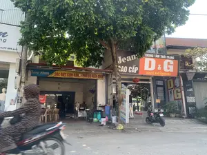 Bức ảnh chụp một con phố với nhiều cửa hàng kinh doanh. Ở tiền cảnh, một người mặc áo chống nắng kẻ sọc đang điều khiển xe máy di chuyển trên đường. Bên phải là vỉa hè với một cây xanh lớn, phía dưới là các cửa hàng.。Có một quán ăn với tấm bạt màu xanh và vàng ghi dòng chữ 'Thắng Nhung' và 'ĐẶC BIỆT CƠM RANG - PHỞ BÒ NAM ĐỊNH'. Bên trong quán có thể thấy bàn ghế cho khách và khu vực bếp với các bình ga và dụng cụ nấu ăn.。Ngay cạnh đó là một trung tâm thời trang lớn với biển hiệu 'Jean CAO CẤP' và biển hiệu màu cam nổi bật 'TRUNG TÂM THỜI TRANG D&G DOLCE AND GABBANA'. Phía trước cửa hàng có các ma-nơ-canh trưng bày quần áo và một chiếc đèn lồng màu đỏ. Một chiếc xe máy khác đang đỗ trên vỉa hè trước cửa hàng.。Các chi tiết văn bản khác có thể đọc được bao gồm số nhà '44' và '46', một biển hiệu dọc có chữ 'TRANG', một biển quảng cáo đứng trên vỉa hè cũng quảng cáo cho 'PHỞ BÒ ĐỊNH' và 'CƠM RANG', và một biển hiệu ghi số điện thoại '0379 899 468'.。（日本語訳）。写真は、多くの商店が並ぶ通りを写したものです。前景には、縞模様の日焼け防止ジャケットを着た人がバイクを運転して道路を移動しています。右側には大きな緑の木がある歩道があり、その下には店が並んでいます。「Thắng Nhung」と「ĐẶC BIỆT CƠM RANG - PHỞ BÒ NAM ĐỊNH」（特製チャーハン - ナムディン風牛肉フォー）と書かれた青と黄色のタープがある飲食店があります。店内には客用のテーブルと椅子、ガスボンベや調理器具のある厨房エリアが見えます。そのすぐ隣には、「Jean CAO CẤP」（高級ジーンズ）の看板と、「TRUNG TÂM THỜI TRANG D&G DOLCE AND GABBANA」（D&G ドルチェ＆ガッバーナ ファッションセンター）という目立つオレンジ色の看板を掲げた大きなファッションセンターがあります。店の前には服を展示しているマネキンと赤い提灯があります。店の前の歩道には別のバイクが駐車しています。その他読み取れる文字情報には、番地の「44」と「46」、縦長の看板の「TRANG」という文字、歩道に置かれた「PHỞ BÒ ĐỊNH」（ナムディン風牛肉フォー）と「CƠM RANG」（チャーハン）を宣伝する立て看板、そして電話番号「0379 899 468」を記した看板が含まれます。