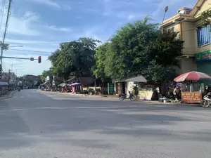 Bức ảnh chụp một con đường nhựa tại một khu vực dân cư, có thể là một thị trấn ở Việt Nam, vào một ngày nắng đẹp, trời xanh. Bên phải đường là một khu chợ nhỏ tự phát với nhiều quầy hàng rong bán đồ ăn. Có những người bán hàng đang hoạt động, một người đội nón đang sắp xếp hàng hóa dưới một chiếc ô lớn. Các phương tiện di chuyển chủ yếu là xe máy. Một cột đèn tín hiệu giao thông đang hiển thị đèn đỏ với đồng hồ đếm ngược chỉ số 14. Dọc hai bên đường là những ngôi nhà nhiều tầng và cây xanh lớn. Các dòng chữ có thể đọc được trên các biển hiệu bao gồm: 1. Biển hiệu quầy thuốc màu xanh lá: 'QUẦY THUỐC MINH KHANG, CHUYÊN THUỐC NỘI, NGOẠI, THỰC PHẨM CHỨC NĂNG, SỮA BỘT Y TẾ'. 2. Biển hiệu trên xe đẩy bán vịt quay: 'BÙI MƯỜI VỊT QUAY, LỢN QUAY MÁ MẬT, NHẬN QUAY ĐÁM TIỆC, ĐT: 09.7 813229'. 3. Biển hiệu trên một cửa hàng: 'QUÝ TÍN ĐIỂM, DC: THỊ T...'.。（日本語訳）。写真は、晴れた青空の日、ベトナムの町と思われる住宅街のアスファルト道路を撮影したものです。道路の右側には、食べ物を売る多くの露店が並ぶ、自然発生的な小さな市場があります。商売をしている売り手がおり、一人はノンラーをかぶり、大きなパラソルの下で商品を並べています。主な交通手段はバイクです。交通信号機が赤信号を表示しており、カウントダウンタイマーは14を示しています。道路の両脇には、多層階の家々と大きな緑の木々が並んでいます。看板で読み取れる文字は以下の通りです：1. 緑色の薬局の看板：「ミンカン薬局、内科・外科の薬、機能性食品、医療用粉ミルク専門」。2. ローストダックを売る屋台の看板：「ブイ・ムオイ ローストダック、マクマット風味のローストポーク、パーティー用のロースト承ります、電話番号：09.7 813229」。3. ある店の看板：「クイ・ティン・ディエム、住所：ティ T...」。