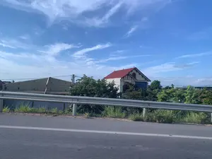 Bức ảnh chụp một khung cảnh ven đường, có thể là ở khu vực ngoại ô hoặc nông thôn. Ở tiền cảnh là một phần của con đường nhựa và một dải hộ lan bằng kim loại. Phía sau dải hộ lan là cây cối xanh tươi. Nổi bật trong khung cảnh là một ngôi nhà hai tầng có tường trắng, mái ngói màu đỏ. Bên cạnh ngôi nhà này là một công trình lớn hơn, có vẻ là một nhà kho hoặc nhà xưởng với mái và tường bằng tôn. Bầu trời trong xanh với những đám mây trắng mỏng. Có thể thấy cột điện và dây điện trong ảnh. Không có người hay hoạt động cụ thể nào được ghi lại, chỉ là một cảnh quan tĩnh được chụp từ một phương tiện đang di chuyển.。（日本語訳）。写真は道端の風景を写しており、郊外か田舎の地域である可能性があります。前景には、アスファルトの道路の一部と金属製のガードレールがあります。ガードレールの後ろには、青々とした木々があります。風景の中で目立っているのは、白い壁と赤い瓦屋根の2階建ての家です。この家の隣には、より大きな建物があり、屋根と壁がトタンでできた倉庫か工場のようです。空は青く澄み渡り、薄い白い雲が浮かんでいます。写真には電柱と電線が見えます。人や特定の活動は記録されておらず、移動中の乗り物から撮影された静的な風景です。