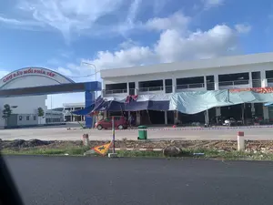 Bức ảnh ghi lại khung cảnh tại một khu chợ lớn, có thể là Chợ đầu mối phía Tây ở Thanh Hóa. Trên cổng vào có một tấm biển lớn hình vòm với dòng chữ màu đỏ "CHỢ ĐẦU MỐI PHÍA TÂY". Phía sau là các tòa nhà chợ lớn, nhiều tầng, sơn màu trắng, có kiến trúc hiện đại. Phía trước một dãy nhà, một số người, có thể là tiểu thương, đang hoạt động dưới những tấm bạt lớn được dựng tạm. Có một chiếc ô tô con màu đỏ và một vài chiếc xe máy đang đỗ. Các hoạt động dường như là buôn bán hoặc sắp xếp hàng hóa. Các vật thể khác có trong ảnh bao gồm một thùng rác màu xanh, dải băng rào chắn sọc đỏ trắng, và một biển báo công trường bị đổ bên lề đường. Khung cảnh lề đường còn ngổn ngang đất đá cho thấy khu vực này có thể đang được xây dựng. Bầu trời trong xanh và có mây.。（日本語訳）。写真は、タインホア省にある西部卸売市場と思われる、ある大きな市場の光景を記録しています。入口のゲートの上には、「CHỢ ĐẦU MỐI PHÍA TÂY」という赤い文字が書かれた大きなアーチ型の看板があります。その後ろには、白く塗られた近代的な建築様式の、大きな多層階の市場の建物があります。一列に並んだ建物の前では、おそらく小規模な商人と思われる数人が、仮設の大きな防水シートの下で活動しています。赤い乗用車1台と数台のバイクが駐車しています。その活動は、商品の売買または整理のようです。写真に写っている他の物には、緑色のゴミ箱、赤と白の縞模様のバリケードテープ、そして道端に倒れた工事現場の標識が含まれます。道端の光景には土や石が散乱しており、この地域が建設中である可能性を示しています。空は青く晴れ渡り、雲があります。