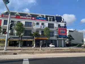 Bức ảnh chụp một dãy nhà phố thương mại nhiều tầng, màu trắng, nằm ven một con đường vào ban ngày. Bầu trời trong xanh, có ít mây. Con đường và vỉa hè phía trước tòa nhà đang trong quá trình thi công, bề mặt còn đất đá.。Các dòng chữ trên các biển hiệu có thể đọc được bao gồm:。- Trên tầng thượng: "GLOBAL ACADEMY", "FACEBOOK: GLOBAL", "...ĐƠN THANH HÓA", "ENGLISH COFFEE", "HOTLINE. 02373 201 555", "SƠN VILAZA", "NHÀ PHÂN PHỐI TRUNG GIANG".。- Biển hiệu dọc bên phải: "SUNTEK DUY THU", "SUNHOUSE", "THƯƠNG HIỆU GIA DỤNG HÀNG ĐẦU VIỆT NAM", "ĐẠI LÝ DUY THU", "ĐC: Đường Nguyễn Chích, khu phố Nam Sơn, TT Rừng Thông, Thanh Hóa", "ĐT: 0345 809 789 - 0385 083 181".。- Tầng trệt: "...NG THÀNH PH... SỐ 47 KHU PHỐ NAM SƠN MÓN ĂN DÂN TỘC - ĐẶC SẢN", "ĐIỂM DỪNG CHÂN LÝ TƯỞNG", "ĐẶC SẢN PHỞ TRÂU TƯƠI", "Coffee NGUYỄN PIAGGIO".。Địa điểm trong ảnh là một khu phố kinh doanh sầm uất, có thể ở Thị trấn Rừng Thông, Thanh Hóa, dựa trên thông tin địa chỉ. Các hoạt động diễn ra bao gồm kinh doanh của các cửa hàng như trung tâm tiếng Anh, quán cà phê, đại lý đồ gia dụng và quán ăn. Một chiếc ô tô con màu trắng đang đỗ trước một cửa hàng. Phía xa bên phải, có một người đội nón lá đang đẩy một chiếc xe thô sơ. Các vật thể nổi bật gồm có tòa nhà nhiều tầng, các biển hiệu quảng cáo, cột đèn giao thông đang bật đèn xanh, cây xanh, và một chiếc ô tô.。（日本語訳）。写真は、日中に撮影された、道路沿いに建つ白くて多層階の商店街の一角を写しています。空は青く澄み渡り、雲はほとんどありません。建物の前の道路と歩道は工事中で、地面はまだ土や石が残っています。看板に書かれている読み取れる文字は以下の通りです：。- 最上階：「GLOBAL ACADEMY」、「FACEBOOK: GLOBAL」、「...ĐƠN THANH HÓA」、「ENGLISH COFFEE」、「HOTLINE. 02373 201 555」、「SƠN VILAZA」、「NHÀ PHÂN PHỐI TRUNG GIANG」。- 右側の縦看板：「SUNTEK DUY THU」、「SUNHOUSE」、「ベトナムのトップ家庭用品ブランド」、「ĐẠI LÝ DUY THU」、「住所：タインホア省、ルン・トン町、ナムソン街区、グエン・チク通り」、「電話：0345 809 789 - 0385 083 181」。- 1階：「...NG THÀNH PH... ナムソン街区47番地 民族料理 - 特産品」、「理想的な休憩所」、「特産品 新鮮な水牛のフォー」、「Coffee NGUYỄN PIAGGIO」。写真の場所は、住所情報に基づくと、タインホア省ルン・トン町にある可能性のある、にぎやかな商業地区です。ここで行われている活動には、英語センター、カフェ、家庭用品代理店、飲食店などの店舗の営業が含まれます。ある店の前に白い乗用車が1台駐車しています。遠くの右側には、ノンラーをかぶった人が手押し車を押しています。目立つ物としては、多層階の建物、広告看板、青信号が点灯している信号機、緑の木々、そして一台の自動車があります。