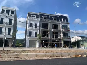 Bức ảnh chụp một công trường xây dựng với các dãy nhà phố liền kề đang trong các giai đoạn hoàn thiện khác nhau. Ở trung tâm là một ngôi nhà đang trong quá trình xây dựng, để lộ kết cấu bê tông thô và chưa có cửa sổ. Phía trước ngôi nhà này có một giàn giáo lớn làm bằng tre. Bên trái là một ngôi nhà gần như đã hoàn thiện với lớp sơn trắng, cửa cuốn ở tầng trệt và ban công sắt. Có một vài công nhân đang làm việc tại công trường. Phía trước các tòa nhà là một con đường nhựa rộng rãi. Bầu trời trong xanh, có mây trắng, cho thấy thời tiết đẹp. Xung quanh có các cột đèn đường, dây điện và một vài cây xanh mới trồng.。（日本語訳）。この写真は、隣接するタウンハウスの列がさまざまな完成段階にある建設現場を写したものです。中央には建設中の家があり、むき出しのコンクリート構造が見え、まだ窓がありません。この家の前には、竹で作られた大きな足場があります。左側には、白い塗装が施され、1階にシャッター、鉄製のバルコニーが付いた、ほぼ完成した家があります。現場では数人の作業員が働いています。建物の前には、広々としたアスファルトの道路があります。空は青く澄み渡り、白い雲が浮かんでおり、天気が良いことを示しています。周囲には街灯の柱、電線、そして新しく植えられた数本の木があります。