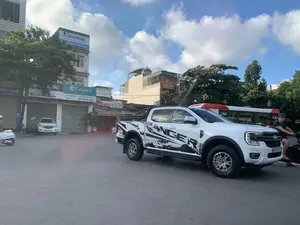 Bối cảnh là một con đường phố tại thành phố Thanh Hóa, Việt Nam, với các phương tiện giao thông đang di chuyển. Nổi bật ở trung tâm là một chiếc xe bán tải Ford Ranger màu trắng có dán đề can chữ 'RANGER' lớn ở bên hông. Bên phải chiếc xe bán tải là một người đàn ông đang điều khiển xe máy. Ở phía sau có thể thấy một phần của chiếc xe buýt màu đỏ và trắng, và ở bên trái là một chiếc xe tay ga màu trắng. Dọc hai bên đường là những tòa nhà cao tầng, nơi có nhiều cửa hàng kinh doanh. Có thể đọc được nhiều biển hiệu quảng cáo, bao gồm:。- Biển hiệu của nhà phân phối Đào Hòa cho các hãng điện máy: 'DAIKIN - CHUYÊN GIA ĐIỀU HÒA KHÔNG KHÍ HÀNG ĐẦU NHẬT BẢN', 'CÔNG TY TNHH SẢN XUẤT & THƯƠNG MẠI ĐÔNG Á - NHÀ PHÂN PHỐI ĐÀO HÒA', và biển hiệu 'Casper - A BRAND FROM THAILAND' tại địa chỉ '76B'. Một biển hiệu khác có ghi địa chỉ 'Phú Sơn - TP. Thanh Hóa'.。- Biển hiệu quán ăn: 'Thanh Thảo - CƠM BÌNH DÂN' và 'CƠM BÌNH DÂN - BIA HƠI'.。- Một biển hiệu khác ghi 'QUẤN THẮNG' cùng số điện thoại 'ĐT: 0948 989 925 - 0941 260 33'.。Bầu trời trong xanh, có mây trắng.。（日本語訳）。背景はベトナムのタインホア市にある街路で、交通車両が移動しています。中央で目立っているのは、側面に大きな「RANGER」の文字のデカールが貼られた白いフォード・レンジャーのピックアップトラックです。ピックアップトラックの右側には、バイクを運転している男性がいます。後方には赤と白のバスの一部が見え、左側には白いスクーターがあります。道路の両側には高層ビルが立ち並び、多くの店舗が入っています。多くの広告看板が読み取れます。以下が含まれます：。- 家電メーカーの代理店ダオホアの看板：「DAIKIN - 日本のトップ空調専門家」、「ドンアー製造・貿易有限会社 - ダオホア代理店」、そして住所「76B」にある「Casper - A BRAND FROM THAILAND」の看板。別の看板には「フーソン - タインホア市」という住所が書かれています。- 食堂の看板：「タイン・タオ - 大衆食堂」と「大衆食堂 - 生ビール」。- 別の看板には「QUẤN THẮNG」と電話番号「ĐT: 0948 989 925 - 0941 260 33」が書かれています。空は青く澄んでおり、白い雲があります。