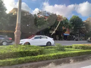 Bức ảnh chụp cảnh một con đường đô thị ở Việt Nam, nhìn từ bên trong một chiếc xe đang chạy. Có nhiều phương tiện đang lưu thông trên đường, nổi bật là một chiếc ô tô sedan màu trắng đang di chuyển. Ngoài ra còn có một chiếc ô tô màu đen, một chiếc xe bán tải đang đỗ và một người đi xe máy. Con đường có dải phân cách ở giữa với hàng rào cây xanh được cắt tỉa. Dọc hai bên đường là các tòa nhà nhiều tầng, một phần bị che khuất bởi cây cối. Bầu trời trong xanh và có mây. Có một số biển hiệu của các cửa hàng, nhưng do ảnh bị nhòe vì chuyển động nên chỉ đọc được một phần. Một biển hiệu màu vàng có chữ '...MOTO... CAFE...'. Một biển hiệu màu xanh lá cây có vẻ là 'TIỆM CẮT TÓC'. Một biển hiệu lớn hơn màu cam có chữ 'Đại lý...' nhưng không thể đọc rõ chi tiết.。（日本語訳）。ベトナムの都市部の道路の風景を、走行中の車内から撮影した写真です。道路上には多くの車両が通行しており、走行中の白いセダン車が目立っています。その他にも、黒い自動車、駐車中のピックアップトラック、バイクに乗った人がいます。道路の中央には、刈り込まれた植木の柵がある中央分離帯があります。道路の両脇には高層ビルが立ち並び、その一部は木々で隠されています。空は青く晴れており、雲があります。いくつかの店舗の看板がありますが、写真が動きでブレているため、一部しか読み取れません。黄色い看板には「...MOTO... CAFE...」という文字があります。緑色の看板は「TIỆM CẮT TÓC」（理髪店）のようです。より大きなオレンジ色の看板には「Đại lý...」（代理店...）という文字がありますが、詳細ははっきりと読み取れません。