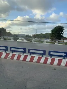 Bức ảnh chụp cảnh quan từ một phương tiện đang di chuyển. Ở tiền cảnh là mặt đường nhựa và một dải phân cách bê tông được sơn sọc đỏ và trắng. Trên dải phân cách có đặt các biển báo chỉ hướng màu xanh lam với mũi tên màu trắng chỉ về bên phải. Phía sau là một cây cầu bắc qua một con sông. Lan can cầu có các trụ bê tông màu trắng với hoa văn trang trí màu vàng. Phía xa, bên kia sông, có thể thấy cây cối, nhà cửa, cột điện và một ngọn đồi. Bầu trời có nhiều mây nhưng vẫn có những khoảng xanh.。（日本語訳）。移動中の乗り物から撮影された風景写真です。前景には、アスファルトの路面と、赤と白の縞模様に塗装されたコンクリートの中央分離帯があります。中央分離帯の上には、右を指す白い矢印が付いた青い方向指示標識が設置されています。その後ろには、川にかかる橋があります。橋の欄干には、黄色い装飾模様が施された白いコンクリートの柱があります。遠くの川の向こう側には、木々、家々、電柱、そして丘が見えます。空は雲が多いですが、まだ青い部分もあります。