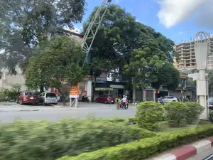 Đây là một khung cảnh đường phố tại một khu vực đô thị ở Việt Nam, được chụp từ một phương tiện đang di chuyển. Dọc bên đường là các tòa nhà, cửa hàng và cây xanh lớn. Có người dân đang đi xe máy trên đường. Một số ô tô được đỗ ở lề đường. Ở phía sau, một tòa nhà cao tầng đang trong quá trình xây dựng. Có nhiều biển hiệu với các dòng chữ có thể nhận dạng được, bao gồm: "Viettel", "HÀNG SẮT SƯ THÀNH", "...NG TRÌNH XÂY DỰNG", và các biển quảng cáo "SALE", "SALE HẾT CỠ", "SỐC CHƯA TỪNG CÓ", "LED". Phía trước là dải phân cách có cây bụi và lề đường được sơn vạch đỏ trắng.。（日本語訳）。これはベトナムのある都市部の街の風景で、移動中の乗り物から撮影されたものです。道沿いには建物、店、そして大きな緑の木々があります。人々がバイクに乗って道路を走っています。何台かの車が路肩に駐車されています。後方には、高層ビルが建設中です。認識できる文字が書かれた多くの看板があり、それには「Viettel」、「HÀNG SẮT SƯ THÀNH」、「...NG TRÌNH XÂY DỰNG」、そして「SALE」、「SALE HẾT CỠ」、「SỐC CHƯA TỪNG CÓ」、「LED」といった広告看板が含まれます。手前には低木のある中央分離帯と、赤と白の線で塗装された路肩があります。