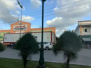 Hình ảnh ghi lại quang cảnh bên ngoài một tòa nhà lớn màu cam, đây là Chợ đầu mối Nông lâm thủy sản Đông Hương. Trên biển hiệu của tòa nhà có dòng chữ lớn màu đỏ 'CHỢ ĐẦU MỐI' và dòng chữ nhỏ hơn 'NÔNG LÂM THỦY SẢN ĐÔNG HƯƠNG'. Phía trước tòa nhà, một chiếc xe rơ moóc đang đỗ, chở một thùng container lớn màu trắng. Trên thùng container có các ký hiệu như 'CGMU 503211 1', '45R1' và một chữ 'M' lớn. Ngoài ra còn có một vài chiếc ô tô và xe máy đang đỗ trên con đường. Bối cảnh cho thấy hoạt động có thể liên quan đến việc vận chuyển và tập kết hàng hóa. Bầu trời trong xanh, có mây và nhiều đường dây điện.。（日本語訳）。画像は、大きなオレンジ色の建物の外の光景を記録したもので、これはドンフオン農林水産卸売市場です。建物の看板には、大きな赤い文字で「CHỢ ĐẦU MỐI」と、それより小さい文字で「NÔNG LÂM THỦY SẢN ĐÔNG HƯƠNG」と書かれています。建物の前には、トレーラートラックが1台駐車しており、大きな白いコンテナを積んでいます。コンテナには、「CGMU 503211 1」、「45R1」といった記号と、大きな「M」の文字があります。さらに、道路には数台の自動車とバイクも駐車しています。この背景は、商品の輸送や集荷に関連する活動が行われている可能性を示しています。空は青く澄み、雲があり、多くの電線があります。