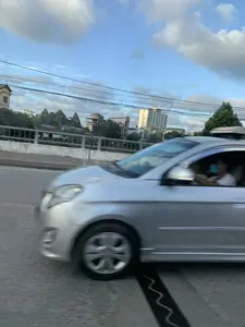 Bức ảnh được chụp từ một phương tiện đang di chuyển, ghi lại cảnh quan giao thông đô thị trên một cây cầu. Ở cận cảnh, một chiếc ô tô màu bạc đang chạy trên đường, hình ảnh bị nhòe do chuyển động. Bên trong xe có thể thấy người ngồi, trong đó một người ở ghế phụ đang đeo khẩu trang. Phía sau lan can cầu là khung cảnh thành phố với nhiều tòa nhà có kiến trúc và chiều cao khác nhau, bao gồm cả một tòa nhà chung cư cao tầng. Cây cối mọc xen kẽ giữa các công trình xây dựng. Bầu trời trong xanh, có mây trắng và nhiều đường dây điện chằng chịt ở phía trên. Trên mặt đường có một khe co giãn của cầu được sơn vạch đen hình lượn sóng.。（日本語訳）。この写真は移動中の乗り物から撮影されたもので、橋の上の都市交通の風景を記録しています。手前には、銀色の自動車が道路を走行しており、その像は動きによってぼやけています。車内には人が座っているのが見え、そのうち助手席にいる一人はマスクを着用しています。橋の欄干の後ろには、高層マンションを含む、建築様式や高さが異なる多くの建物がある街の風景が広がっています。建設物の間に木々が点在して生えています。空は青く澄んでおり、白い雲があり、頭上には多くの電線が張り巡らされています。路面には、波形の黒い線で塗装された橋の伸縮継手があります。