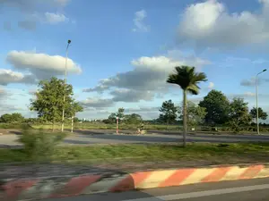 Bức ảnh được chụp từ một phương tiện đang di chuyển, ghi lại cảnh quan ven một con đường lớn ở khu vực ngoại ô hoặc nông thôn. Phía trước là một dải phân cách hoặc lề đường được sơn sọc đỏ và trắng, bị mờ do chuyển động. Có một con đường nhựa chạy song song, trên đó có một người đang đi xe đạp. Dọc theo con đường là các cột đèn đường, cây xanh, bao gồm cả một cây cọ bị nhòe hình. Phía xa là những cánh đồng và một vài công trình xây dựng. Bầu trời trong xanh và có nhiều mây trắng. Không có chữ viết nào có thể đọc được trong ảnh.。（日本語訳）。この写真は移動中の乗り物から撮影されたもので、郊外または田舎にある大きな道路沿いの風景を捉えています。前方には、赤と白の縞模様に塗られた中央分離帯または路肩があり、動きによってぼやけています。平行して走るアスファルトの道があり、その上を一人、自転車に乗っている人がいます。道に沿って街灯の柱や緑の木々があり、その中には像がぼやけたヤシの木も一本含まれています。遠くには田畑といくつかの建設物があります。空は青く澄んでおり、白い雲が多く浮かんでいます。写真の中には、読み取れる文字はありません。