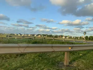 Bức ảnh ghi lại quang cảnh vùng nông thôn ở Thanh Hóa, được chụp từ một phương tiện đang di chuyển. Ở phía trước là một dải hộ lan bằng kim loại chạy dọc theo con đường. Phía sau hàng rào hộ lan là một con đường nhựa, xa hơn là những cánh đồng xanh và một khu dân cư với nhiều nhà cửa, cây cối. Có thể thấy các cột điện cao thế và cột thu phát sóng viễn thông ở phía xa. Bên phải có một biển báo giao thông hình tròn trên cột sọc đỏ trắng, nhưng không rõ nội dung. Bầu trời trong xanh và có nhiều mây.。（日本語訳）。この写真はタインホア省の田園風景を記録したもので、移動中の乗り物から撮影された。手前には、道路に沿って続く金属製のガードレールがある。ガードレールの後ろにはアスファルトの道路があり、その先には緑の田畑と、多くの家や木々がある住宅地が広がっている。右側には赤と白の縞模様のポールに円形の交通標識があるが、内容は不明瞭である。空は青く澄んでおり、多くの雲がある。