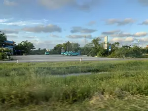 Bối cảnh là một khu vực ven đường, có thể là một trạm dừng nghỉ hoặc trạm xăng. Ở phía sau, có một bãi đỗ xe rộng rãi, nơi một số chiếc ô tô điện VinFast màu xanh lam đang đỗ, trông giống như xe taxi. Có một biển báo lớn màu xanh và trắng ghi các thông tin: 'LÀNG RỒNG SƠN', 'E5 RON 92', 'DIESEL 0.05S', 'DẦU BÔI TRƠN'. Ngay bên cạnh là một biển báo nhỏ hơn có logo 'VINFAST' và biểu tượng trạm sạc xe điện. Xung quanh khu vực này có nhiều cây xanh, cột đèn đường và một cột điện cao thế ở phía xa. Tiền cảnh là một cánh đồng cỏ xanh và một con mương nhỏ, hình ảnh ở khu vực này bị mờ do chuyển động, cho thấy ảnh được chụp từ một phương tiện đang di chuyển. Bầu trời trong xanh và có nhiều mây.。（日本語訳）。背景は道端のエリアで、休憩所かガソリンスタンドのようです。後方には広々とした駐車場があり、そこには数台の青いビンファスト（VinFast）製電気自動車が駐車しており、タクシーのように見えます。青と白の大きな看板があり、「LÀNG RỒNG SƠN」、「E5 RON 92」、「DIESEL 0.05S」、「DẦU BÔI TRƠN（潤滑油）」という情報が書かれています。そのすぐ隣には、「VINFAST」のロゴと電気自動車充電ステーションのシンボルが描かれた、より小さな看板があります。このエリアの周りには多くの緑の木々、街灯、そして遠くには高圧電線の鉄塔があります。前景には緑の草地と小さな水路があり、この部分の画像は動きによってぼやけており、移動中の乗り物から撮影されたことを示しています。空は青く澄んでおり、雲が多く浮かんでいます。