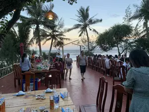 Bối cảnh là một nhà hàng ngoài trời ven biển vào lúc hoàng hôn. Rất đông người đang tụ tập ăn tối tại các dãy bàn ghế gỗ dài. Mọi người đang ăn uống, trò chuyện vui vẻ trong không khí nhộn nhịp. Phía sau là bãi biển với sóng vỗ, được bao quanh bởi nhiều cây dừa và cây phi lao. Không gian được trang trí bằng một chiếc đèn lồng làm bằng mây tre đan và các dây đèn nhỏ. Trên bàn ăn có nhiều bát đĩa, cốc, chai nước khoáng hiệu 'Aquafina' và lon nước ngọt màu cam hiệu 'Splash'. Gần đó có một thùng nhựa màu xanh dương có chữ 'VINAMILK'.。（日本語訳）。背景は、夕暮れ時の海辺にある屋外レストランです。大勢の人々が、長い木製のテーブルと椅子の列で夕食のために集まっています。人々は賑やかな雰囲気の中、飲食をしたり、楽しくおしゃべりをしたりしています。後ろには波が打ち寄せるビーチがあり、多くのヤシの木とモクマオウの木に囲まれています。空間は、籐や竹で編まれたランタンと小さなストリングライトで飾られています。食卓の上には、多くの皿や椀、コップ、「アクアフィナ」ブランドのミネラルウォーターのボトル、そして「スプラッシュ」ブランドのオレンジ色の炭酸飲料の缶があります。その近くには、「VINAMILK」という文字が入った青いプラスチック製のコンテナがあります。