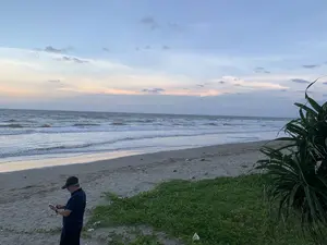 Bối cảnh là một bãi biển vào lúc hoàng hôn hoặc bình minh. Bầu trời có mây và ánh sáng dịu nhẹ ở đường chân trời. Biển có nhiều sóng vỗ vào bờ cát. Ở phía bên trái của bức ảnh, một người đàn ông lớn tuổi mặc áo phông màu xanh đậm, quần tối màu và đội mũ lưỡi trai màu đen đang đứng trên cát và sử dụng điện thoại thông minh. Phía bên phải có thảm thực vật ven biển, bao gồm cây rau muống biển và một bụi cây dứa dại. Không có thông tin dạng chữ viết nào trong ảnh.。（日本語訳）。背景は、夕暮れ時または夜明け時のビーチです。空は曇っており、地平線には柔らかな光があります。海には多くの波が砂浜に打ち寄せています。写真の左側には、濃い青色のTシャツ、暗い色のズボンを着用し、黒い野球帽をかぶった年配の男性が砂の上に立ち、スマートフォンを使用しています。右側には、グンバイヒルガオやアダンの茂みなど、海岸の植生があります。写真には文字情報はありません。