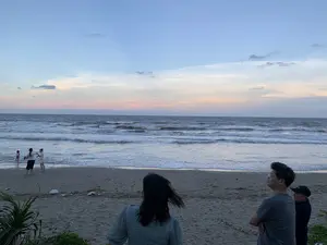 Đây là một khung cảnh trên bãi biển vào lúc hoàng hôn hoặc bình minh. Bầu trời có màu xanh lam pha lẫn sắc hồng ở đường chân trời. Dưới mặt biển, những con sóng đang vỗ vào bờ cát. Có một nhóm người đang có mặt trên bãi biển. Ở phía trước, một vài người đang đứng ngắm cảnh biển. Ở phía xa hơn, gần mép nước, có hai người lớn và một em bé đang chơi đùa trong sóng. Bãi biển có cát và một ít cây cỏ mọc ở gần bờ. Không có văn bản hay chữ viết nào có thể nhận dạng được trong ảnh.。（日本語訳）。これは夕暮れ時または夜明け時のビーチの光景です。空は青く、地平線にはピンク色が混じっています。海では、波が砂浜に打ち寄せています。ビーチには一群の人々がいます。手前には、数人が海の景色を眺めて立っています。もっと遠く、水際に近いところでは、大人2人と赤ちゃん1人が波の中で遊んでいます。ビーチは砂浜で、岸の近くにはいくつかの草木が生えています。写真の中には、認識できるテキストや文字はありません。