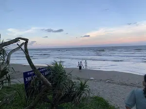 Bối cảnh là một bãi biển vào lúc hoàng hôn hoặc bình minh. Bầu trời có màu xanh lam chuyển dần sang hồng và cam ở đường chân trời. Biển có sóng vỗ vào bờ cát.。Ở phía trước bên trái, có một tấm biển báo màu xanh dương với chữ trắng, bị che khuất một phần bởi cây cối và một thân cây khô. Các dòng chữ có thể nhận dạng được là 'ỦY BAN N...', 'XÃ QUẢNG...', và các chữ lớn 'KHU V...'. Bên dưới có chữ 'Y HIỂM', có thể là một phần của từ 'NGUY HIỂM'. Đây có thể là biển cảnh báo của Ủy ban Nhân dân một xã ven biển.。Trên bãi biển, có một nhóm người đang đi dạo. Một người phụ nữ mặc áo sơ mi trắng và váy đen đang đi trên cát, dắt tay một đứa trẻ nhỏ. Một người khác mặc trang phục màu trắng đang đi ở mép nước. Ở ngoài khơi xa, có thể thấy một vài vật thể nổi màu cam, có thể là phao hoặc cờ báo hiệu. Ở góc dưới bên phải của bức ảnh, một phần lưng của một người phụ nữ mặc áo hoa màu xanh lam đang nhìn ra biển.。（日本語訳）。背景は、夕暮れ時または夜明け時のビーチです。空は青色で、地平線に向かって徐々にピンクとオレンジ色に変わっています。海には波が砂浜に打ち寄せています。左手前には、青地に白文字の看板があり、木々と一本の枯れ木によって部分的に隠されています。認識できる文字は「ỦY BAN N...」、「XÃ QUẢNG...」、そして大きな文字の「KHU V...」です。その下には「Y HIỂM」という文字があり、これは「NGUY HIỂM」（危険）という単語の一部である可能性があります。これは、ある沿岸の社の人民委員会による警告看板である可能性があります。ビーチでは、一団の人々が散歩しています。白いシャツと黒いスカートを着た女性が、幼い子供の手を引いて砂の上を歩いています。白い服を着た別の人が水際を歩いています。遠くの沖合には、オレンジ色の浮遊物がいくつか見えますが、それはブイか信号旗かもしれません。写真の右下隅には、青い花柄の服を着た女性の背中の一部が写っており、海を見ています。