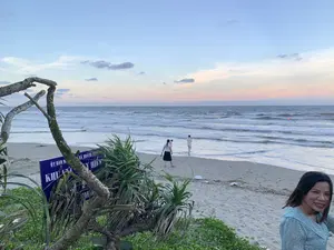 Bức ảnh chụp cảnh một bãi biển vào lúc hoàng hôn hoặc bình minh. Bầu trời có màu xanh và hồng nhạt. Ở phía trước bên phải, một người phụ nữ mặc váy hoa màu xanh nhạt đang mỉm cười nhìn vào máy ảnh. Ở trung tâm, một vài người đang đi dạo trên bãi cát và lội nước; một phụ nữ đang bế một em bé. Biển có sóng và có thể thấy một vài chiếc phao màu cam ở phía xa. Ở phía trước bên trái, có một tấm biển báo màu xanh bị che khuất một phần bởi cành cây và cây dứa dại. Các dòng chữ có thể đọc được trên biển báo là 'ỦY BAN N...', 'XÃ QUẢNG TH...', và chữ lớn 'KHU VỰC...'. Bãi cát có cây rau muống biển mọc.。（日本語訳）。写真は、夕暮れ時または夜明け時のビーチの光景を撮影したものです。空は青色と薄いピンク色をしています。右手前には、水色の花柄のワンピースを着た女性がカメラに向かって微笑んでいます。中央では、数人が砂浜を散歩したり、水の中を歩いたりしています。一人の女性が赤ちゃんを抱いています。海には波があり、遠くにオレンジ色のブイがいくつか見えます。左手前には、木の枝とアダンの木によって一部が隠れている青い看板があります。看板で読み取れる文字は、「ỦY BAN N...」、「XÃ QUẢNG TH...」、そして大きな文字で「KHU VỰC...」です。砂浜にはグンバイヒルガオが生えています。