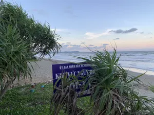 Bức ảnh chụp cảnh một bãi biển vào lúc hoàng hôn hoặc bình minh. Ở phía trước, có một tấm biển báo màu xanh dương với chữ trắng, bị che khuất một phần bởi những bụi cây dứa dại. Nội dung trên biển báo có thể đọc được là 'ỦY BAN NHÂN DÂN XÃ QUẢNG NHAM' và dòng chữ lớn hơn bên dưới có thể là 'KHU VỰC NGUY HIỂM...'. Phần còn lại của văn bản không thể nhìn rõ. Khung cảnh cho thấy một bãi cát trải dài ra biển, với những con sóng đang vỗ vào bờ. Phía xa trên mặt biển là hình ảnh một hòn đảo hoặc một dãy núi. Bầu trời có màu sắc nhẹ nhàng với vài gợn mây. Không có người trong ảnh, khung cảnh rất yên tĩnh.。（日本語訳）。夕暮れか夜明けのビーチの風景を写した写真です。手前には、白い文字が書かれた青い看板があり、アダンの茂みによって一部が隠されています。看板に書かれた内容は「ỦY BAN NHÂN DÂN XÃ QUẢNG NHAM」と読み取れ、その下にあるより大きな文字は「KHU VỰC NGUY HIỂM...」かもしれません。文字の残りの部分ははっきりと見えません。風景には、海へと広がる砂浜と、岸に打ち寄せる波が見えます。遠くの海上には、島または山脈の姿が見えます。空は優しい色合いで、いくつかの薄い雲があります。写真には人がおらず、風景はとても静かです。