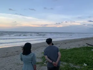 Cảnh chụp tại một bãi biển vào lúc hoàng hôn hoặc bình minh. Có hai người, một nam và một nữ, đang đứng trên bãi cát và nhìn ra biển. Họ được chụp từ phía sau. Người phụ nữ ở bên trái mặc một chiếc váy màu xanh nhạt có hoa văn. Người đàn ông ở bên phải mặc áo phông màu xám, chắp tay sau lưng. Bầu trời có mây với màu sắc của buổi chạng vạng. Biển có sóng đang vỗ vào bờ. Trên bãi cát có một ít thảm thực vật màu xanh lá cây, có thể là rau muống biển, và một ít rác.。（日本語訳）。夕暮れか夜明けのビーチで撮影された風景。男性と女性の二人が砂浜に立ち、海を眺めている。彼らは後ろから撮影されている。左側の女性は花柄の薄い青色のワンピースを着ている。右側の男性は灰色のTシャツを着て、手を後ろで組んでいる。空は曇っており、夕暮れ時の色合いである。海には波があり、岸に打ち寄せている。砂浜には、おそらくグンバイヒルガオと思われる緑色の植物が少しと、ゴミが少しある。