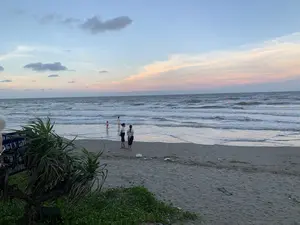 Quang cảnh tại một bãi biển ở Thanh Hóa vào thời điểm hoàng hôn. Bầu trời có những dải mây màu hồng và cam trên nền xanh nhạt. Dưới mặt nước, sóng biển đang vỗ vào bờ cát. Có một vài người đang có mặt tại bãi biển: hai người phụ nữ đang đứng trên cát và nhìn ra biển, một em bé mặc đồ bơi màu hồng đang chơi ở vùng nước nông, và một người khác đứng ở phía xa hơn. Hoạt động của họ là thư giãn và tắm biển. Ở ngoài khơi, có thể thấy những chiếc phao hoặc cờ màu cam. Ở góc dưới bên trái của ảnh, có một bụi cây và một tấm biển màu xanh. Dù bị che khuất, có thể nhận ra các từ như '...AN XÃ...' và 'NGUY HIỂM', cho thấy đây là một biển cảnh báo.。（日本語訳）。タインホア省のあるビーチの夕暮れ時の光景。空には薄青色の背景にピンクとオレンジ色の帯状の雲がある。水面の下では、海の波が砂浜に打ち寄せている。ビーチには数人の人がいる：2人の女性が砂の上に立って海を眺めており、ピンク色の水着を着た小さな子供が浅瀬で遊んでおり、もう一人がさらに遠くに立っている。彼らの活動はリラックスと海水浴である。写真の左下隅には、茂みと青い看板がある。隠れてはいるが、「...AN XÃ...」や「NGUY HIỂM」（危険）といった言葉が認識でき、これが警告看板であることを示している。