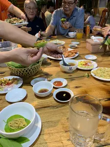 Bối cảnh là một bữa ăn tối, có thể tại một nhà hàng hoặc một buổi tụ họp gia đình. Nhiều người, bao gồm cả những người đàn ông lớn tuổi, đang ngồi quanh một chiếc bàn gỗ lớn. Họ đang cùng nhau thưởng thức một bữa ăn thịnh soạn với nhiều món hải sản.。Trên bàn có các món ăn như tôm nướng, hàu nướng (một người đang bưng đĩa hàu), và một món trông giống gỏi hoặc nộm. Ngoài ra còn có một đĩa đồ ăn kèm gồm xoài xanh, hành tím, và ớt thái lát. Mọi người đang dùng tay để cuốn thức ăn với các loại lá rau sống được đặt trong một chiếc rổ tre.。Các vật dụng trên bàn bao gồm nhiều bát đĩa sứ màu trắng, đũa, thìa, và các bát nhỏ đựng nước chấm như nước tương, tương ớt và nước mắm pha. Có một cốc thủy tinh lớn (có thể là cốc bia), một ly nước cam và một lon nước ngọt. Một trong những chiếc bát sứ có in logo và chữ "QUANG THAI". Mọi người đang ăn uống và trò chuyện vui vẻ.。（日本語訳）。背景は夕食の席で、レストランか家族の集まりかもしれません。年配の男性を含む多くの人々が、大きな木製のテーブルを囲んで座っています。彼らは一緒に、多くの海鮮料理からなる豪華な食事を楽しんでいます。その他にも、青いマンゴー、紫玉ねぎ、スライスした唐辛子からなる付け合わせの皿があります。皆、竹かごに盛られた様々な種類の生の葉野菜で、手を使って食べ物を巻いています。テーブルの上にある物には、多くの白い陶磁器の皿や椀、箸、スプーン、そして醤油、チリソース、調合ヌクマムのようなタレを入れるための小鉢が含まれます。大きなガラスのコップ（ビアグラスかもしれません）、オレンジジュースのグラス一杯、そしてソフトドリンクの缶が一本あります。陶磁器の椀の一つには、「QUANG THAI」というロゴと文字が印刷されています。皆、飲食しながら楽しくおしゃべりしています。