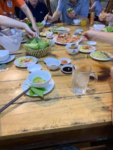 Một nhóm người đang tụ tập ăn uống tại một nhà hàng hoặc quán ăn, ngồi quanh một chiếc bàn gỗ. Trên bàn bày biện nhiều món ăn, nổi bật là một đĩa tôm nướng, một rổ rau thơm và các loại lá để cuốn, cùng nhiều bát nhỏ đựng các loại nước chấm khác nhau. Ngoài ra còn có các món ăn kèm như gỏi. Mọi người đang dùng đũa và tay để thưởng thức bữa ăn. Trên bàn còn có xô đá, cốc bia lớn và lon nước ngọt. Một chiếc bát có in logo chữ 'QUANG TRUNG', có thể là tên của nhà hàng.。（日本語訳）。一群の人々がレストランか食堂で集まって飲食しており、木製のテーブルを囲んで座っています。テーブルの上には多くの料理が並べられており、目立つのは焼きエビの皿、香草と巻くための様々な葉物が入った籠、そして様々な種類のつけダレが入った多くの小鉢です。その他に、ゴイ（和え物）のような副菜もあります。みんな箸と手を使って食事を楽しんでいます。テーブルの上には氷の入ったバケツ、大きなビアジョッキ、そしてソフトドリンクの缶もあります。「QUANG TRUNG」という文字のロゴが印刷された椀が一つあり、それはレストランの名前かもしれません。