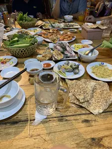 Bức ảnh chụp một bàn ăn thịnh soạn tại một nhà hàng hoặc quán ăn, có thể là một quán hải sản. Một nhóm người đang quây quần ăn uống trong một không khí vui vẻ. Trên bàn gỗ mộc bày rất nhiều món ăn, chủ yếu là hải sản, bao gồm tôm nướng, hàu nướng mỡ hành, mực hấp, và ốc luộc. Ngoài ra còn có các món ăn kèm như bánh đa nướng rắc vừng, hai giỏ rau sống, và nhiều loại nước chấm khác nhau đựng trong các bát nhỏ. Trên bàn còn có bát đĩa, đũa, một cốc bia thủy tinh lớn còn đá, một hộp giấy ăn, một lon nước ngọt hiệu "Sprite" và một chai nước "Aquafina". Một số món ăn đã được dùng dở, cho thấy bữa tiệc đang diễn ra.。（日本語訳）。写真は、レストランか食堂、おそらくは海鮮料理店での豪華な食卓を写しています。一団の人々が楽しい雰囲気の中で集まって飲食しています。素朴な木製のテーブルの上には、主に海鮮料理である多くの料理が並べられており、焼きエビ、ネギ油をかけた焼き牡蠣、蒸しイカ、茹で貝などが含まれています。その他にも、ゴマを振りかけた焼きライスペーパー、生野菜の籠二つ、そして小さな器に入れられた様々な種類のつけダレといった付け合わせ料理があります。テーブルの上にはさらに、皿や椀、箸、氷の入った大きなガラスのビアジョッキ、ティッシュペーパーの箱、「スプライト」ブランドの炭酸飲料の缶、そして「アクアフィーナ」の水のボトルがあります。いくつかの料理は食べかけで、宴会が進行中であることを示しています。