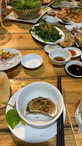 Hình ảnh ghi lại một bữa ăn thịnh soạn trên bàn gỗ, có vẻ là một bữa tiệc hải sản. Trên bàn có rất nhiều món ăn Việt Nam. Ở gần nhất là một chiếc bát trắng đựng một vỏ hàu đã chế biến, bên trong có thịt hàu cùng với hành phi. Xung quanh là các món ăn khác bao gồm một đĩa rau muống xào tỏi, một rổ rau sống, và nhiều đĩa hải sản như tôm nướng và hàu nướng. Ngoài ra còn có nhiều bát nước chấm nhỏ đựng nước tương, tương ớt, nước mắm pha ớt và muối. Một đĩa gỏi nhỏ với xoài, ớt và hành tím cũng được nhìn thấy. Khung cảnh cho thấy một buổi tụ tập ăn uống tại một nhà hàng hoặc tại nhà.。（日本語訳）。画像は木製のテーブルの上の豪華な食事を記録しており、海鮮パーティーのようです。テーブルの上にはたくさんのベトナム料理があります。最も手前には、調理された牡蠣の殻が入った白い器があり、中には牡蠣の身とフライドオニオンが入っています。周りには、ニンニク炒めの空心菜、生野菜のバスケット、そして焼きエビや焼き牡蠣などの多くの海鮮料理を含む他の料理があります。その他にも、醤油、チリソース、唐辛子入りヌクマム、塩が入った多くの小皿のつけダレがあります。マンゴー、唐辛子、紫玉ねぎを使った小さな和え物（ゴイ）の皿も見られます。この光景は、レストランまたは自宅での食事会を示しています。