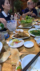 Bối cảnh là một bữa ăn tối tại một nhà hàng hoặc quán ăn, có thể là không gian ngoài trời. Một nhóm người đang ngồi quanh một chiếc bàn gỗ, cùng nhau ăn uống và trò chuyện. Trên bàn bày nhiều món ăn Việt Nam, bao gồm một đĩa cá rán nguyên con, một đĩa rau muống xào, một đĩa gỏi trông giống gỏi tôm với sả, ớt và hành tím, cùng các rổ rau thơm ăn kèm. Ngoài ra còn có bánh đa nướng, vỏ hàu, bát đũa và các chén nước chấm. Một người đang rót đồ uống từ chai màu xanh vào một tô đá lớn. Một người đàn ông lớn tuổi đeo kính đang cầm một con tôm. Về đồ uống, có các lon nước ngọt hiệu 'Mirinda Splash' và chai nước khoáng 'AQUAFINA'. Trên một số đĩa ăn có logo 'QUANG THAI'.。（日本語訳）。背景はレストランか食堂での夕食で、屋外の空間かもしれません。あるグループが木製のテーブルを囲んで座り、一緒に食事をしながらおしゃべりをしています。テーブルの上には、丸ごと一匹の揚げ魚の皿、空心菜の炒め物の皿、レモングラス、唐辛子、紫玉ねぎを使ったエビの和え物のようなサラダの皿、そして付け合わせの香草の籠など、多くのベトナム料理が並べられています。その他にも、焼いたライスペーパー、牡蠣の殻、お椀と箸、そしてつけだれの小鉢があります。一人の人が緑色のボトルから大きな氷の入ったボウルに飲み物を注いでいます。眼鏡をかけた年配の男性がエビを一匹持っています。飲み物については、「ミリンダ・スプラッシュ」ブランドの炭酸飲料の缶と、「アクアフィーナ」のミネラルウォーターのボトルがあります。いくつかの皿には「QUANG THAI」のロゴがあります。