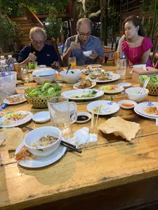 Bối cảnh là một bữa ăn tối tại một nhà hàng hoặc quán ăn có không gian ngoài trời vào buổi tối. Có ba người đang ngồi quanh một chiếc bàn gỗ lớn: hai người đàn ông lớn tuổi và một người phụ nữ. Hai người đàn ông đang dùng bữa, trong khi người phụ nữ mặc áo màu hồng đang ngồi chống cằm. Trên bàn ăn có rất nhiều món ăn, nhiều đĩa đã vơi đi, cho thấy bữa ăn đã diễn ra được một lúc. Các vật thể trên bàn bao gồm: một rổ rau sống, các đĩa hải sản như hàu nướng và tôm, một đĩa gỏi, bánh đa, nhiều bát đĩa sứ màu trắng (một số đĩa có logo màu xanh nhưng không đọc rõ), đũa, thìa, cốc thủy tinh, chai nước lọc, và một lon nước ngọt. Phía sau có cây cối và một cầu thang gỗ.。（日本語訳）。背景は、夜の屋外スペースがあるレストランまたは食堂での夕食です。大きな木製のテーブルの周りに3人が座っています。年配の男性2人と女性1人です。2人の男性は食事をしていますが、ピンク色の服を着た女性は頬杖をついて座っています。食卓の上にはたくさんの料理があり、多くの皿は中身が減っています。これは食事が始まってからしばらく経ったことを示しています。テーブルの上にある物には、生野菜のかご、焼き牡蠣やエビなどの海鮮料理の皿、和え物の皿、バインダー、多くの白い陶磁器の皿や椀（いくつかの皿には青いロゴがありますが、はっきりと読めません）、箸、スプーン、ガラスのコップ、ミネラルウォーターのボトル、そして炭酸飲料の缶が含まれます。後ろには木々と木製の階段があります。