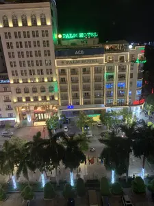 Đây là khung cảnh một góc phố sầm uất vào ban đêm tại một thành phố, được chụp từ trên cao. Các tòa nhà cao tầng được chiếu sáng rực rỡ bằng đèn trang trí và biển hiệu neon. Các văn bản có thể nhận dạng được bao gồm: "PALM HOTEL" (biển hiệu đèn neon xanh lá cây trên nóc nhà và biển hiệu khác trên mặt tiền tòa nhà), "DAI VIET HOTEL", ngân hàng "ACB" (Ngân hàng Á Châu), "SeABank", và một "NHÀ SÁCH" với biển hiệu màu đỏ. Ngoài ra còn có các biển hiệu chỉ nhìn thấy một phần như "THANH... GROUP". Dưới lòng đường, có nhiều ô tô và xe máy đang di chuyển và đậu. Mọi người đang đi bộ trên vỉa hè. Dọc theo con đường là những hàng cây cọ, một số cây được trang trí bằng dây đèn LED. Có một hàng cột cờ treo quốc kỳ Việt Nam.。（日本語訳）。これは、ある都市の夜の賑やかな街角を上から撮影した風景です。高層ビルは装飾灯やネオンサインで煌々と照らされています。認識できる文字には、「PALM HOTEL」（屋上の緑色のネオンサインと建物のファサードにある別の看板）、「DAI VIET HOTEL」、銀行「ACB」（アジア商業銀行）、「SeABank」、そして赤い看板の「NHÀ SÁCH」（書店）が含まれます。その他にも、「THANH... GROUP」のように一部しか見えない看板もあります。道路には、多くの自動車やバイクが走行したり駐車したりしています。人々が歩道を歩いています。道沿いにはヤシの木が並んでおり、そのうちのいくつかはLEDのイルミネーションで飾られています。ベトナム国旗を掲げた旗竿が並んでいます。